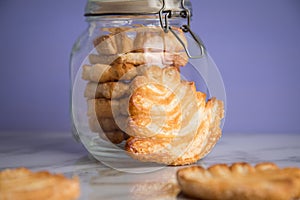 Italian puff pastry fan wavers cookies biscuits with preserving glass jar on marble table and lilac background
