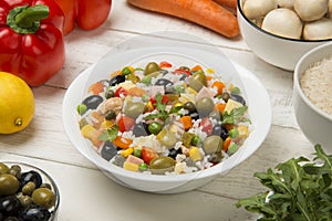 Italian rice salad in a white porcelain salad bowl on a white wooden table with ingredients.