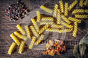 Italian spiral pasta, bay leaves, spices and multi-colored round peppers lie on a textured wooden surface.