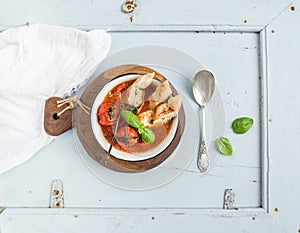 Italian tomato, garlic and basil soup Pappa al Pomodoro in metal bowl with bread on rustic wooden board over light blue