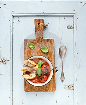 Italian tomato, garlic and basil soup Pappa al Pomodoro in metal bowl with bread on rustic wooden board over light blue