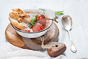 Italian tomato, garlic and basil soup Pappa al Pomodoro in metal bowl with bread on rustic wooden board over light blue