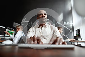 Its all about your money. Focused african male trader sitting by desk and studying analytical reports using pc in the