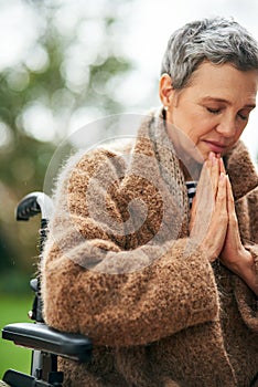 Its faith that keeps her going. a senior woman praying while sitting in her wheelchair outdoors.