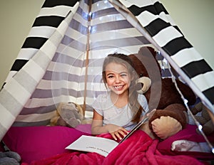 Its her own little reading nook. Portrait of a little girl reading a book with her teddybear in a tent at home.