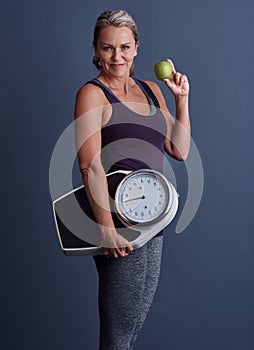 Its a lifestyle. Studio portrait of an attractive mature woman holding an apple and a weightscale against a blue