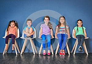 Its our turn now. Studio shot of a diverse group of kids sitting on chairs in a line against a blue background.