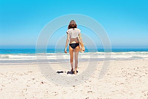 Its about that time for a surf. Rearview shot of a sexy young woman walking towards the ocean with her surfboard.