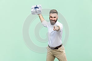 Its for you! Young adult businessman, holding gift box, pointing finger at camera and toothy smile. Isolated on light green backgr