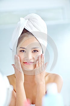 Ive found a skincare routine that works. An attactive young Asian woman applying moisturizer with a towel on her head.