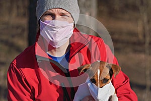 Jack Russell terrier wearing white cotton mouth mask, owner in red jacket with virus face mask in background. Pets are not