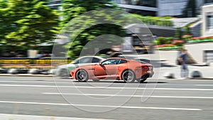 Jaguar F-Type P450 car is driving on the street on high speed. Orange premium sports car in motion
