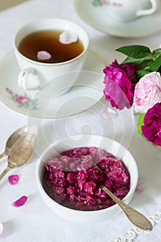 Jam from the petals of the Damascus rose, a cup of green tea and a vase of roses on a light table. Rustic style.