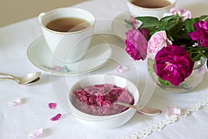 Jam from the petals of the Damascus rose, a cup of green tea and a vase of roses on a light table. Rustic style.