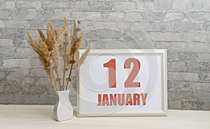 january 12. 12th day of month, calendar date. White vase with ikebana and photo frame with numbers on desktop, opposite