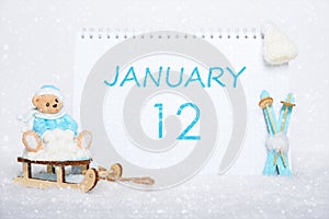January 12th. Teddy bear sitting on a sled, blue skis and a calendar date on white snow. Day 12 of month.