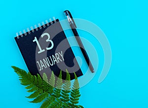 January 13th. Day 13 of month, Calendar date. Black notepad sheet, pen, fern twig, on a blue background