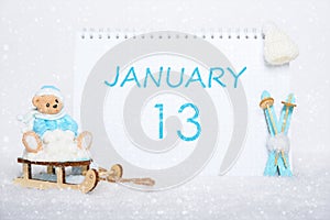 January 13th. Teddy bear sitting on a sled, blue skis and a calendar date on white snow. Day 13 of month.