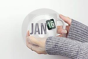 January 16th. Day 16 of month, Calendar date. Closeup of female hands in grey sweater holding cup of tea with month and calendar