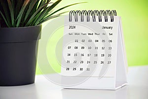 January 2024 desk calendar with potted plant on a desk with green background.