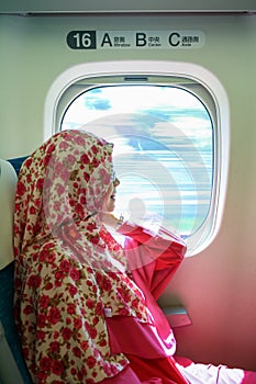 Young asian muslim woman with hijab sitting alone and looking out window smiling inside moving high speed bullet train