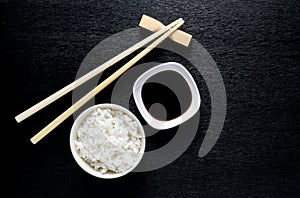 Japanese sushi chopsticks over soy sauce bowl, rice on black