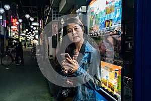 Japanese woman relying on japan vending machines using cellphone relaxing. Young student or female tourist with camera texting sms