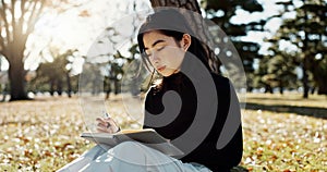 Japanese woman, writing and book in park, thinking and relax by tree, grass or sunshine for peace. Girl, person or