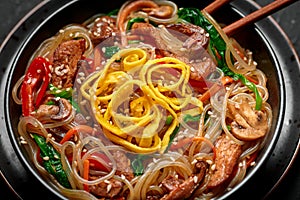 Japchae in black bowl on dark slate table top. Korean cuisine glass chapchae noodles dish with vegetables and meat