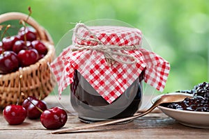 Jar of cherries jam, basket of cherries, saucer and spoon on table