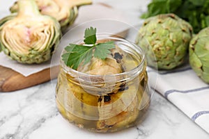 Jar of delicious artichokes pickled in olive oil and fresh vegetables on white marble table