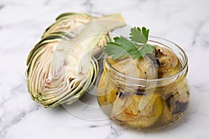 Jar of delicious artichokes pickled in olive oil and fresh vegetables on white marble table