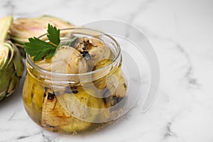 Jar of delicious artichokes pickled in olive oil and fresh vegetables on white marble table, closeup. Space for text