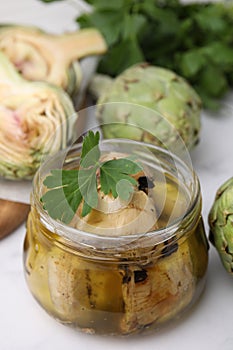 Jar of delicious artichokes pickled in olive oil and fresh vegetables on white table, closeup