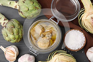 Jar of delicious artichokes pickled in olive oil and ingredients on dark grey wooden table, flat lay