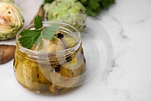 Jar of delicious artichokes pickled in olive oil on white marble table, closeup. Space for text