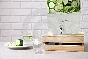 Jar dispenser of fresh cucumber water and glasses on table against brick wall.