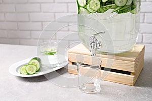 Jar dispenser of fresh cucumber water and glasses on table against brick wall.