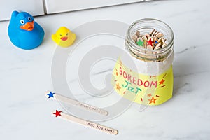 Jar full of wooden sticks with a [BOREDOM JAR] sticker on it on the table near toy ducks