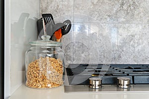 Jar of granola next to modern gas stovetop with kitchen utensils and marble tile backsplace in the backgroud. Bright,