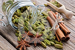 Jar of green cardamom pods. Cinnamon sticks, cardamom seeds, wooden scoop and anise stars on table