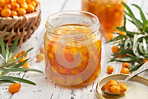 Jar of healthy sea buckthorn jam. Basket of ripe sea buckthorn berries on background. A spoonful of jam in the foreground