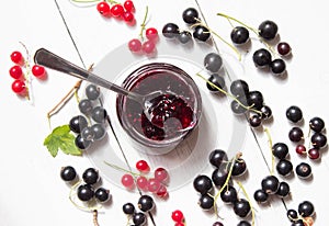Jar of homemade currant jam with spoon. Fresh berries black and red currant on white wooden background. Top view