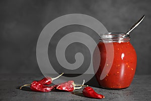 Jar of hot chili sauce with spoon and peppers on table.