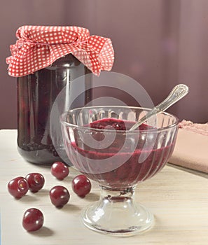 A jar of jam and a vase of jam are on the table, several cherries are scattered around them