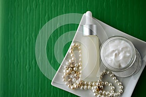 A jar of luxury beauty face cream and serum bottle with pearls on emerald green color background with copy space.