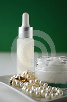 A jar of luxury beauty face cream and serum bottle with pearls on emerald green color background with copy space.