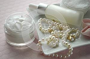 A jar of luxury beauty face cream and serum bottle with pearls on pink color background with copy space.