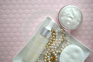 A jar of luxury beauty face cream and serum bottle with pearls on pink color background with copy space.