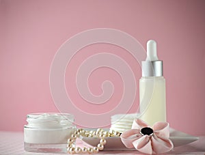 A jar of luxury beauty face cream and serum bottle with pearls on pink color background with copy space.